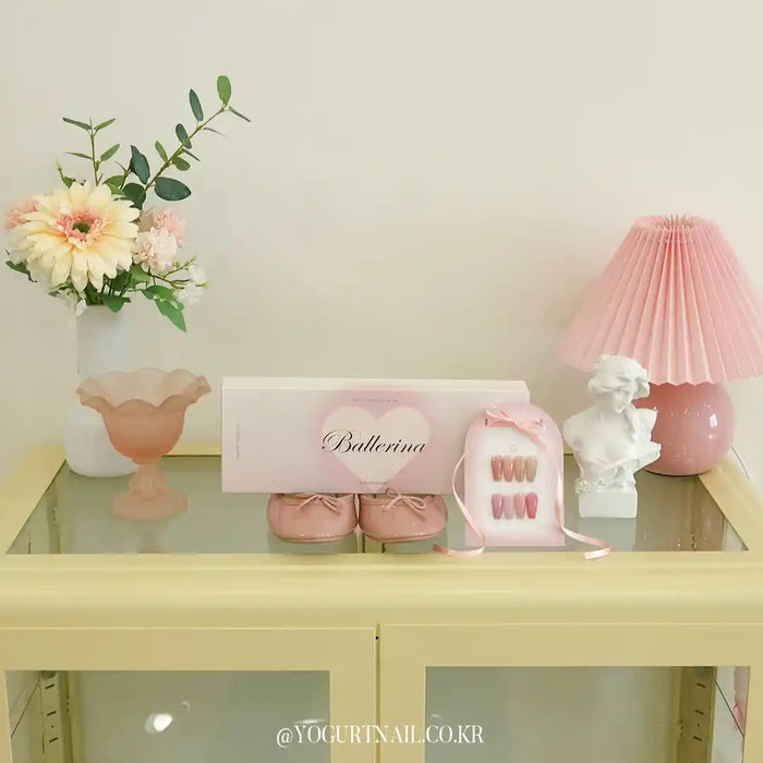 A display of ballet-themed items, including pink ballerina-style shoes, a framed "Ballerina" sign, a pink lamp, and a vase of delicate, pastel-colored flowers. The image evokes a soft, feminine, and elegant aesthetic.