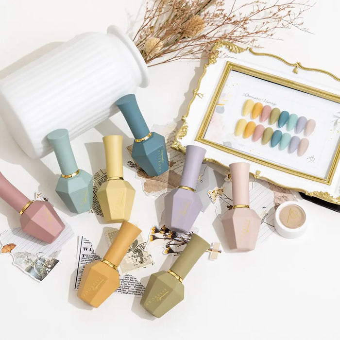 Elegant display of ADORELLE Romantic Vintage nail polish set featuring 8 pastel-colored bottles in matte finishes. Arranged with a framed color swatch, dried flowers, and decorative elements on a white surface, showcasing the collection's soft, vintage-inspired palette and luxurious presentation.