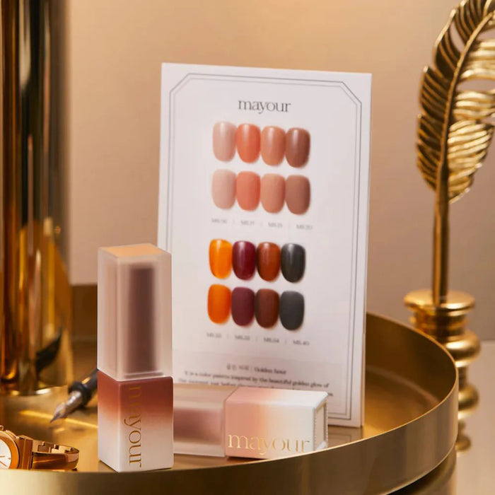 The image shows a product display for the MAYOUR Golden Hour Collection, a set of 8 nail gel colors inspired by the warm and rich tones of the setting sun. The display features a color swatch card showcasing the vibrant shades, along with the product packaging. This elegant and artistic nail polish collection allows customers to transform their manicure routine with mesmerizing nail designs that capture the enchanting beauty of golden hour.