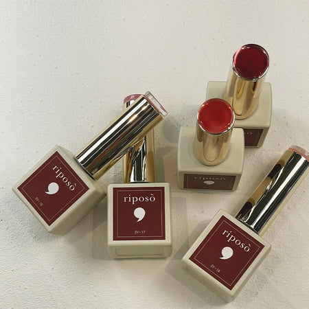 Five highly pigmented, transparent lip glow colors in the Love Love Syrup D Set by Riposo. The set features a range of shades from nude to deep red, with a soft, wet-looking finish that can be intensified by applying multiple coats. The lip products come in sleek, golden packaging to complement the sophisticated, color-changing formula.