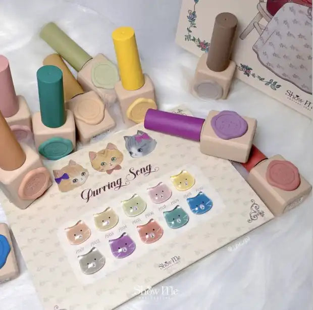 A vibrant and whimsical image showcasing a collection of colorful, cat-themed nail gels from the SHOWME KOREA Purring Song line. The image displays a variety of nail polish bottles, swatches, and other product accessories, all designed with a warm, vintage aesthetic and adorned with cute cat illustrations. This visually appealing product display effectively highlights the brand's signature one-swipe pigmentation and UV/LED curing capabilities, ma