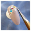 A close-up image of a long, elegant, nude-colored acrylic or gel nail adorned with colorful rhinestones or gems, showcasing an intricate and sophisticated nail art design.