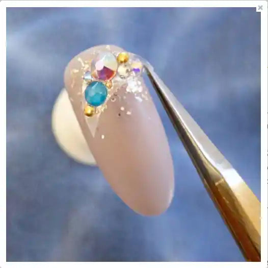 A close-up image of a long, elegant, nude-colored acrylic or gel nail adorned with colorful rhinestones or gems, showcasing an intricate and sophisticated nail art design.