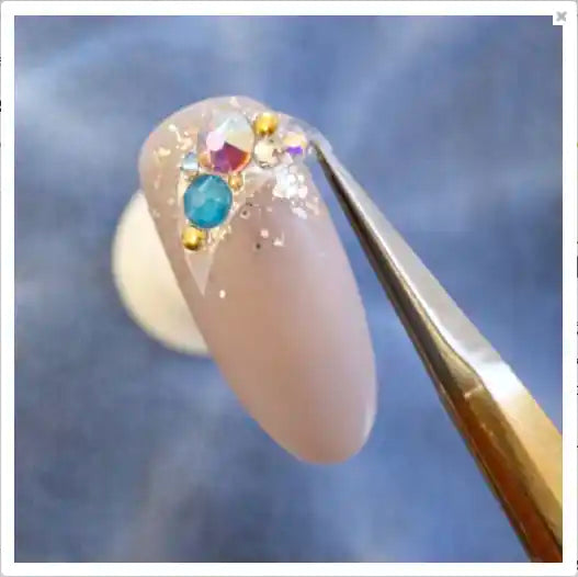 Close-up shot of a textured nail with colorful rhinestones or crystals applied, against a blurred blue sky background. The nail appears to be part of a Tsumekira SP-HBK-104 Yuki Habuka - Plain Sheet Angular Shopify product, showcasing the decorative nail art design.