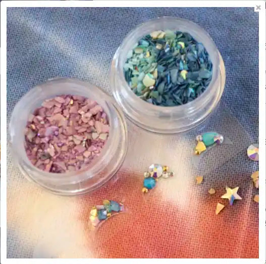 Colorful acrylic nail art decorations in jars, featuring various shapes and sizes of glitter and confetti pieces in shades of green, pink, and gold. The image showcases a wide range of decorative nail art supplies for creating unique and eye-catching nail designs.