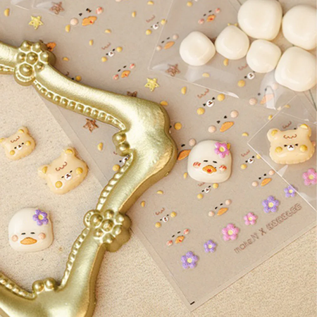 Charming 3D Hoholee and Riri nail charms and stickers adorn a golden nail decor in this whimsical product image, showcasing the playful and adorable designs of the Hoholee x Make.n Charm and Sticker Pack. This visually appealing product shot highlights the unique and delightful nail accessories that can elevate one's nail game with a touch of irresistible cuteness.