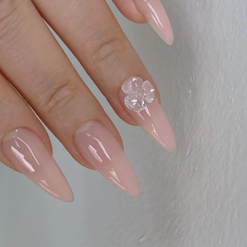 A close-up image of a hand displaying elegant pink manicured nails, one of which features a delicate floral design. The nails appear to be acrylic or gel, with a shiny, polished finish. The image showcases the high-quality craftsmanship and attention to detail of the Nailbayo DOI Aube #02 - 10ml Korean nail product.