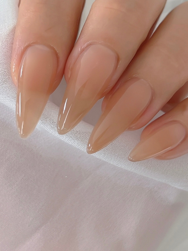 A close-up image of manicured, glossy nude-colored nails with a natural, healthy sheen. The nails are shaped into a pointed, elongated style, creating a sleek and sophisticated look. The image showcases the superior quality and seamless application of the NAILBAYO DOI Nudi Gel Hazelnut product, highlighting its ability to deliver a flawless, long-lasting nail color and finish.