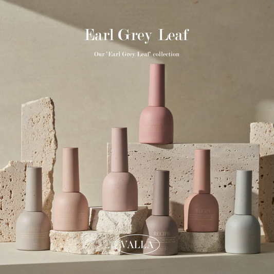 Artistic display of VALLA's Earl Grey Leaf nail polish collection, featuring an assortment of bottles in muted shades of pink, taupe, and grey, artistically arranged on natural stone blocks. The setting uses soft, natural lighting to highlight the smooth, matte finish of the bottles and the sophisticated, earth-toned palette of the collection.